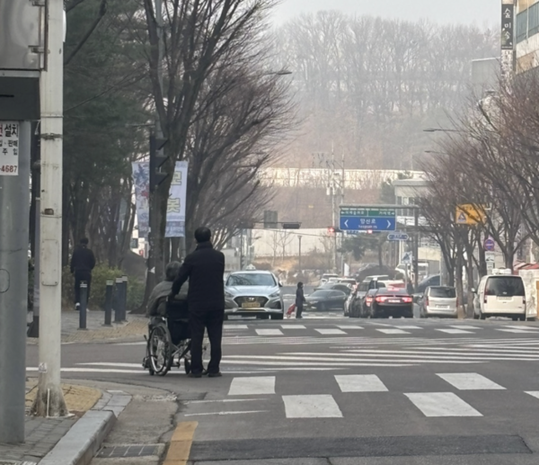 휠체어를 탄 이동약자와 그 보호자가 빈 택시를 잡기 위해 도로에 서 있다. [사진: 석대건 기자]